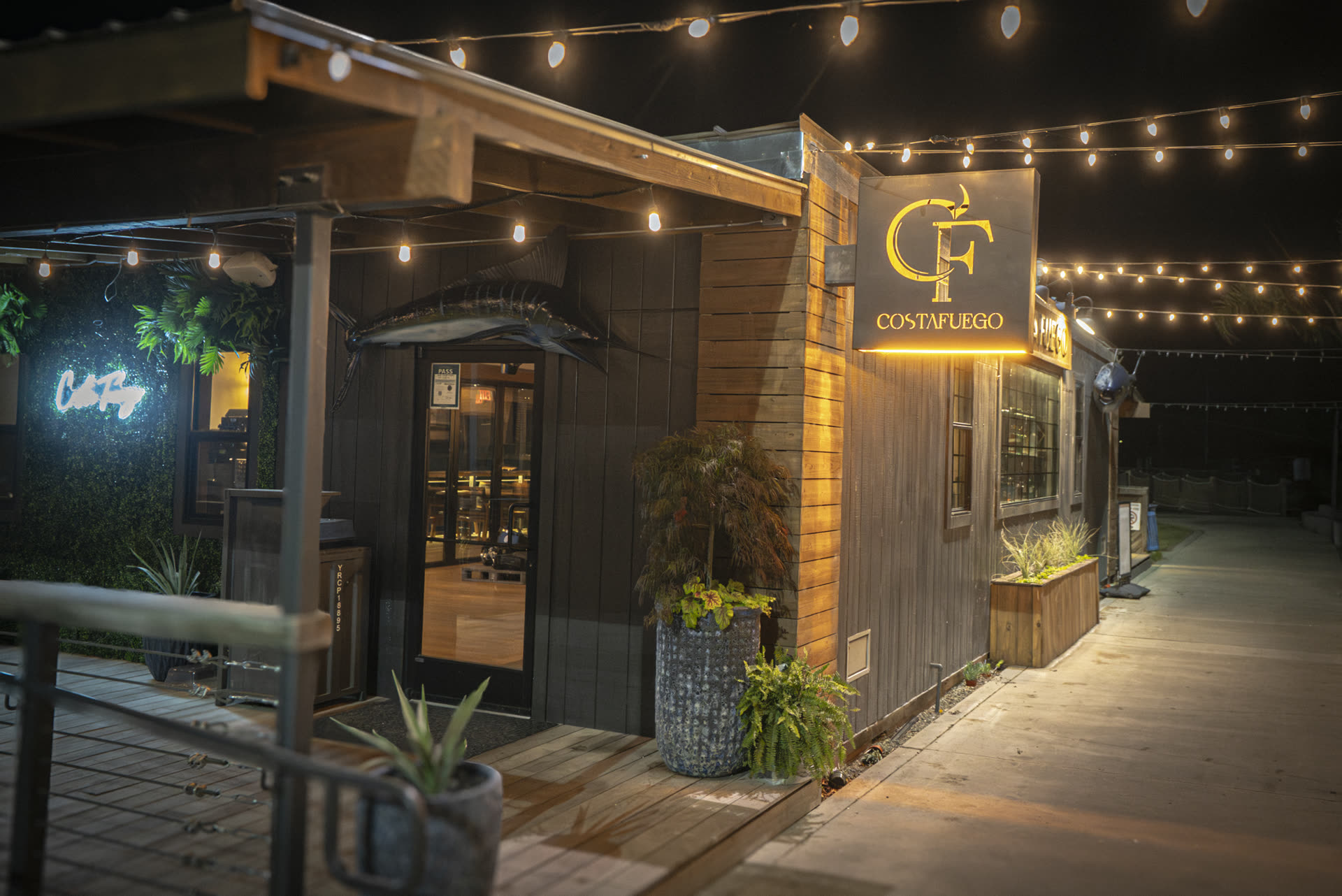 Costa Fuego entrance at night with string lights, the lit CF sign, and the marlin over the door